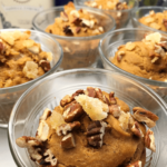 Close-up of five glass bowls filled with pumpkin mousse, each topped with chopped pecans and pieces of candied ginger. In the softly blurred background, hints of Vegan Gluten Free Muffins packaging and greenery are visible.