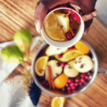 A hand holds a mug of Pear Cider with orange slices and cranberries over a pot filled with fruit slices, cranberries, and cinnamon sticks on a wooden table. Pears and other cozy ingredients are nearby.