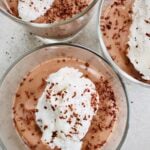 Three glass cups filled with creamy chocolate tofu pudding, each topped with a dollop of whipped cream and sprinkled with chocolate shavings, viewed from above on a light surface.