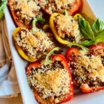 Halved red and yellow Vegan Stuffed Bell Peppers filled with a savory mixture and topped with golden breadcrumbs, arranged in a white baking dish with fresh basil leaves on the side.