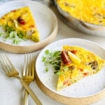 Two plates with slices of Just Egg Frittata, garnished with greens and tomatoes, are set on a light surface next to two gold forks. The remaining frittata is in a pan in the background.