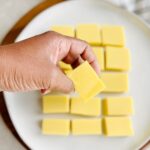 A hand holding a piece of chickpea tofu and a plate of tofu behind