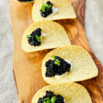 Four potato chips topped with a dollop of vegan sour cream, vegan caviar and chopped green herbs are arranged on a wooden board, with a gold spoon holding more vegan caviar in the background.
