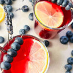 Two glasses of Blueberry Lavender lemonade top with fresh lemon slices and blueberries on table top.