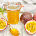 A jar of bright yellow passion fruit jam sits on a marble surface beside whole and halved passion fruits. A gold spoon with jam rests nearby, highlighting its smooth, glossy texture—perfect drizzled with Passion Fruit Syrup for extra flavor.