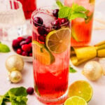A tall glass filled with a red cocktail, ice, lime slices, cranberries and mint sprig, surrounded by fresh limes, cranberries, mint leaves, gold ornaments, and bar tools on a light surface.