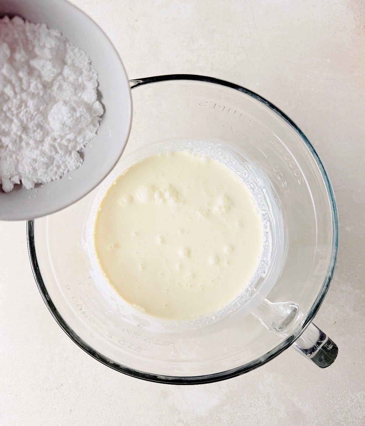 A glass mixing bowl with vegan heavy cream sits on a light surface, ready to become Vegan Lavender Ice Cream. A small white bowl with powdered sugar is held above the creamy mixture, about to be added.