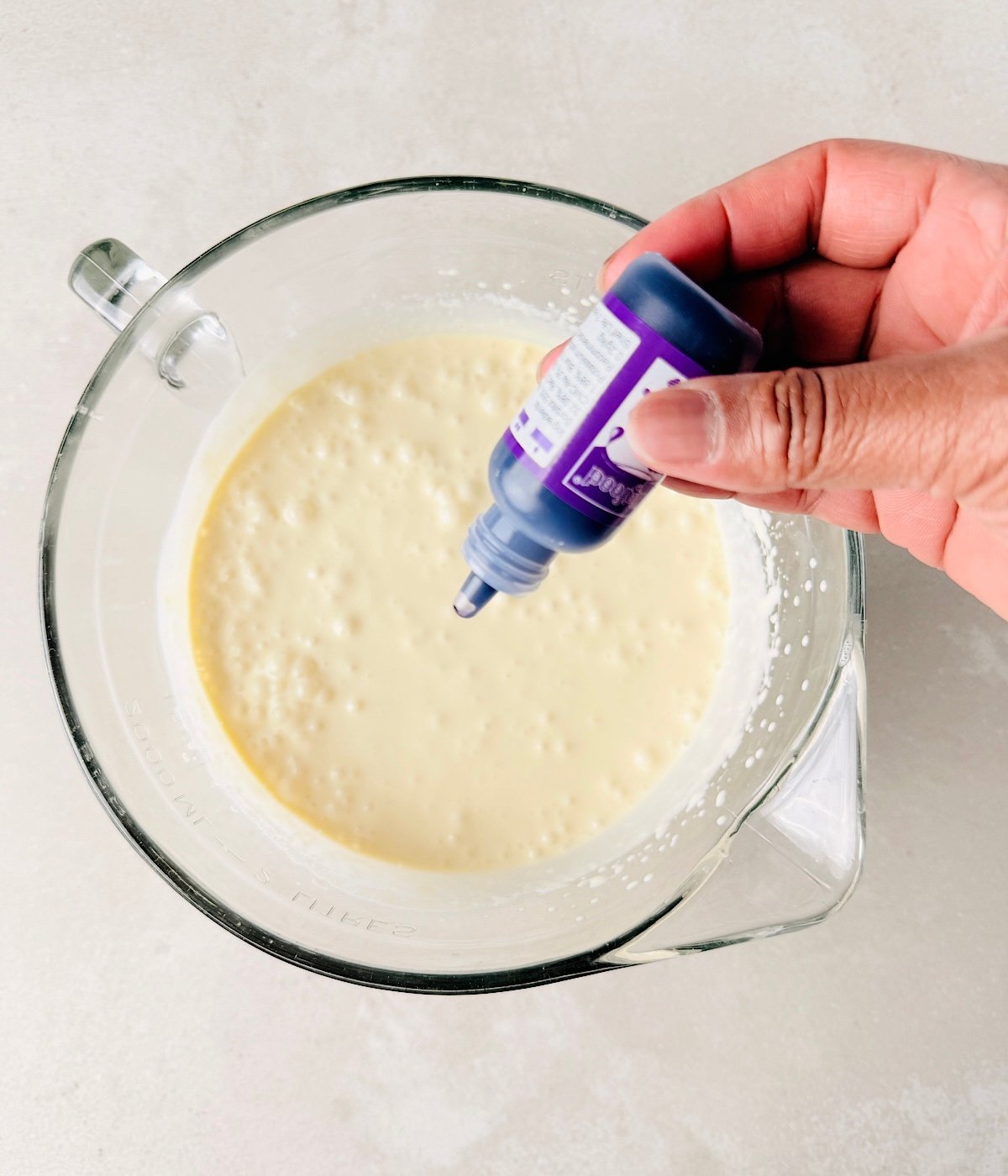 A hand holds a bottle of purple food coloring over a glass mixing bowl filled with pale yellow batter on a light countertop, preparing vegan lavender ice cream.