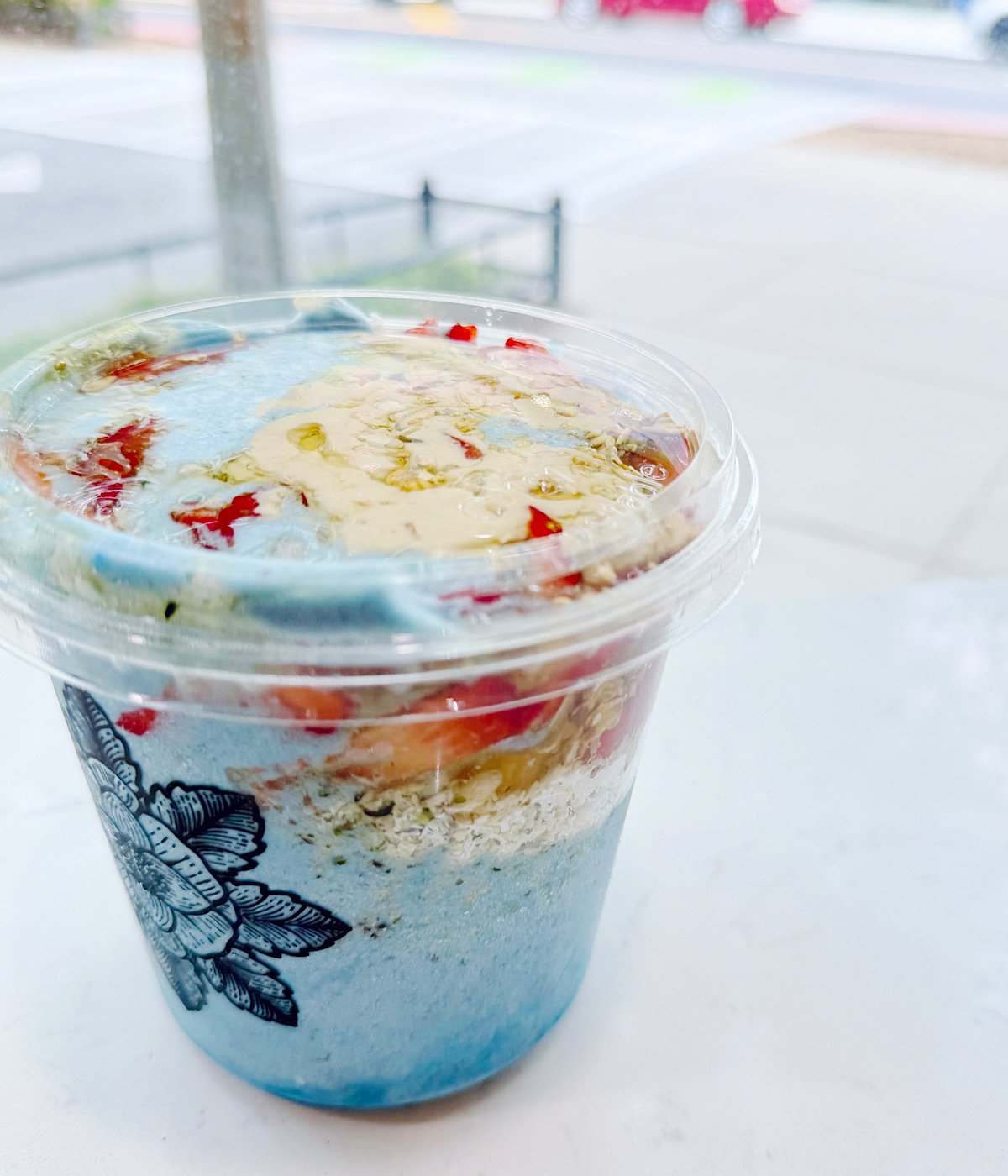 A clear plastic cup filled with a layered blue and white smoothie or parfait, topped with granola, chopped fruits, and drizzled with honey, sits on a white surface near a window.
