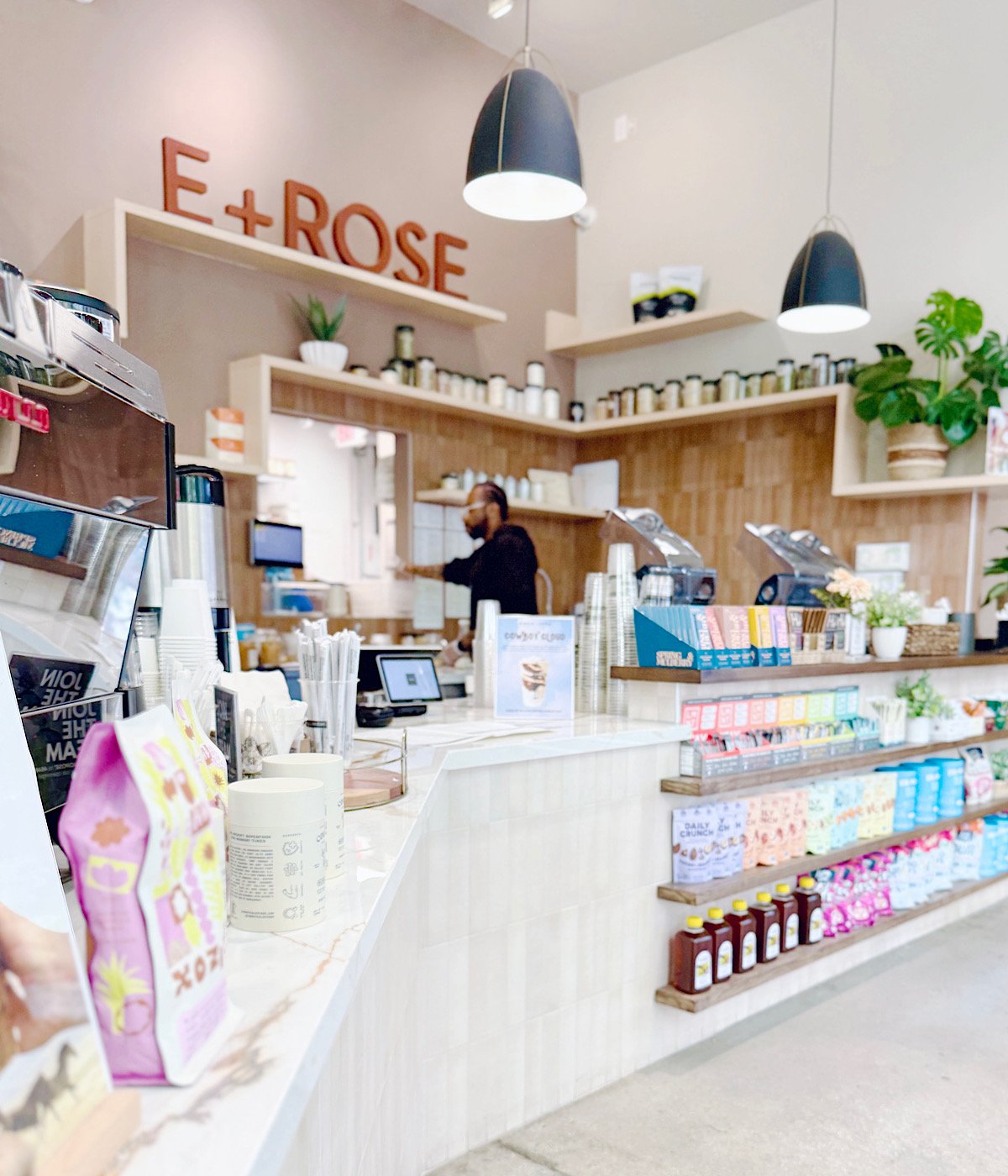 A bright, modern café interior with shelves displaying snacks and drinks, a coffee counter with equipment, and a barista working behind the counter. The sign E + ROSE is visible on the wall.