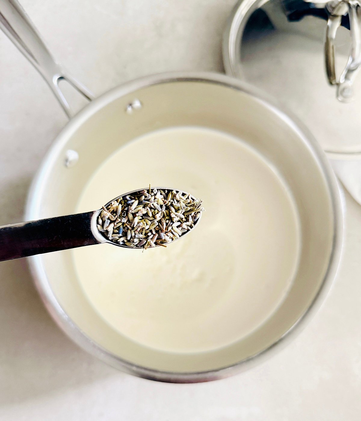 A spoonful of dried lavender buds is held above a saucepan filled with cream or milk, with a metal lid resting beside the pot on a white surface.
