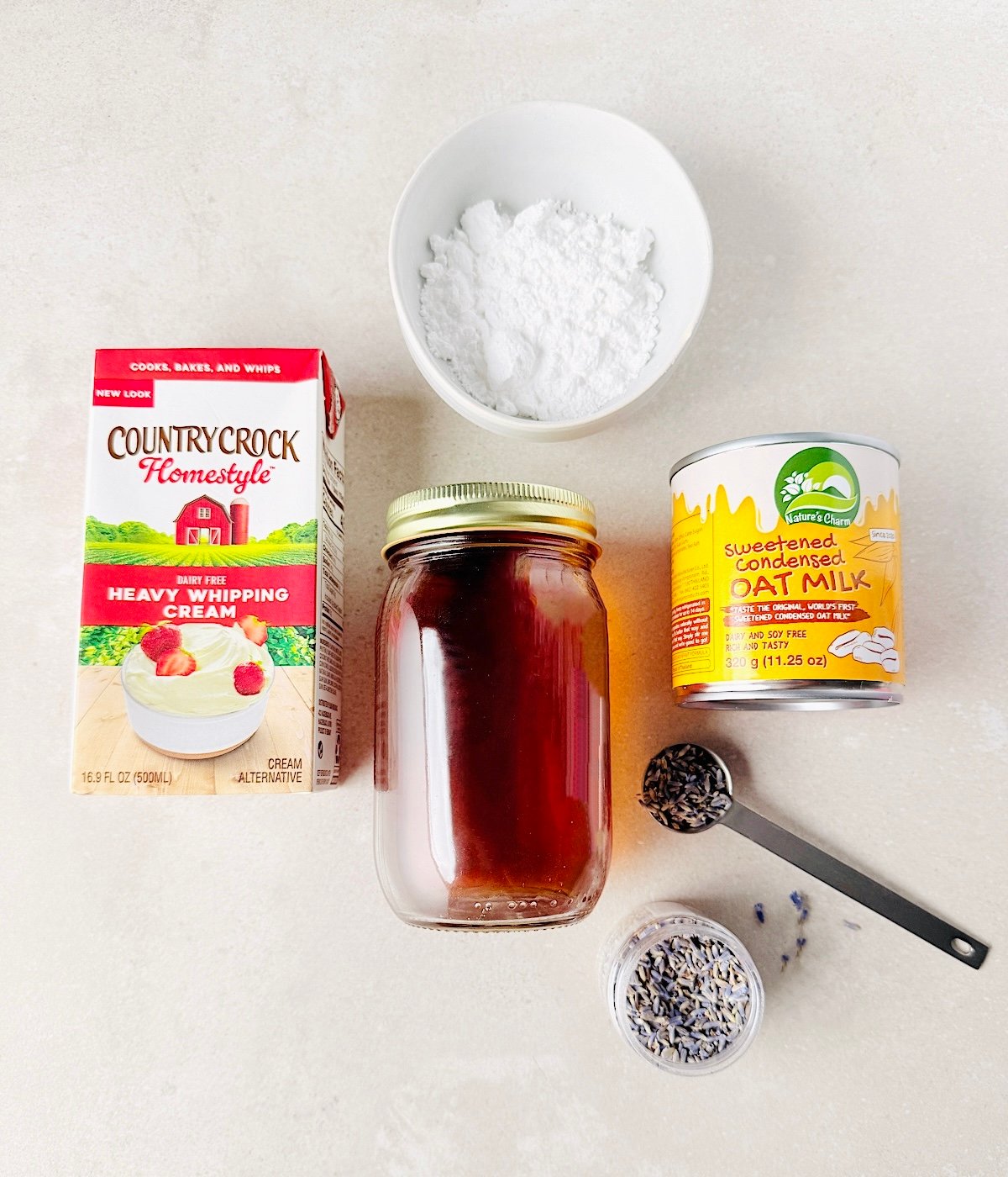 A box of heavy whipping cream, a bowl of powdered sugar, oat milk sweetened condensed milk, lavender extract and dried lavender sit on a white surface—perfect for crafting creamy Vegan Lavender Ice Cream with a floral touch.