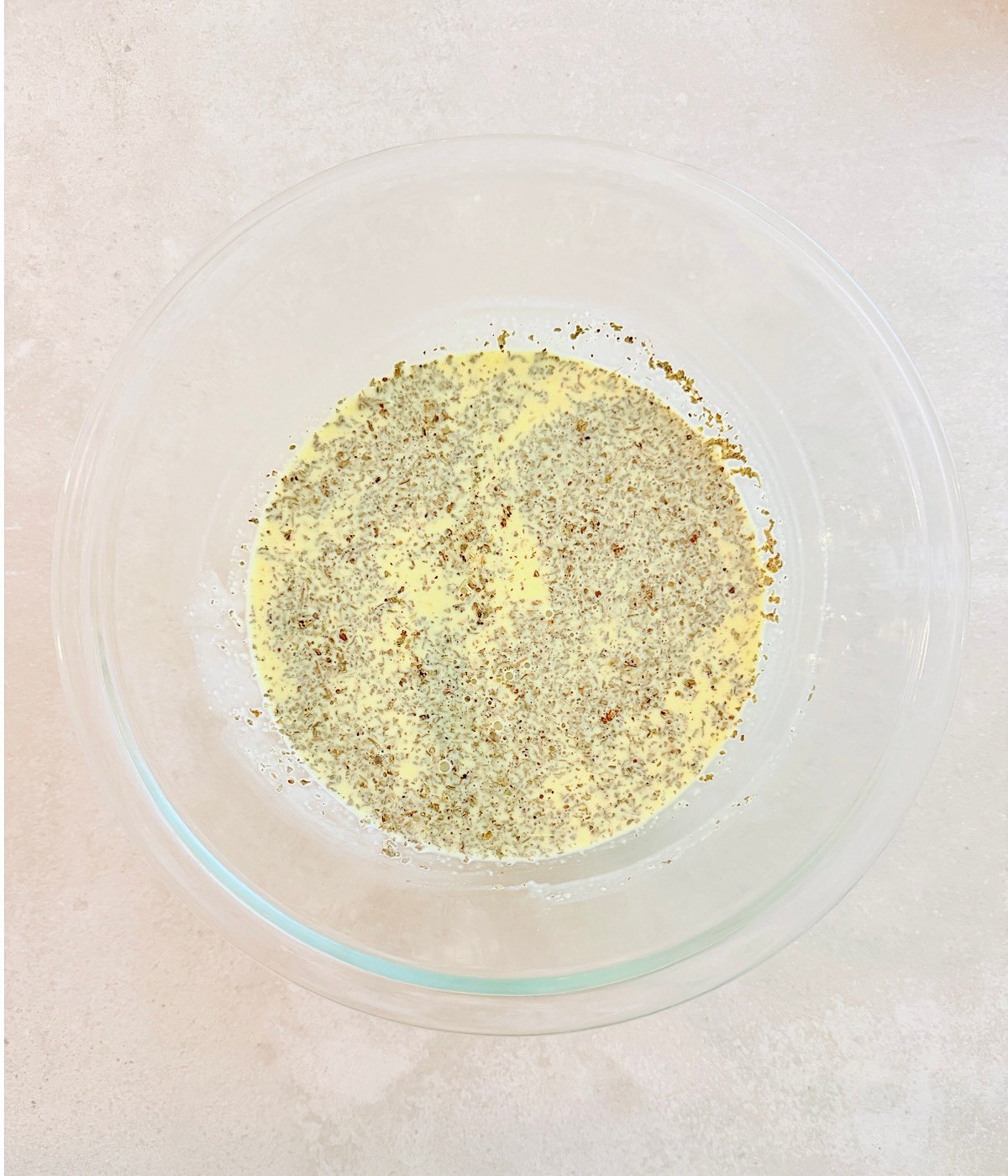 A clear glass bowl with a mixture of beaten vegan eggs and ground black pepper on a light-colored countertop.