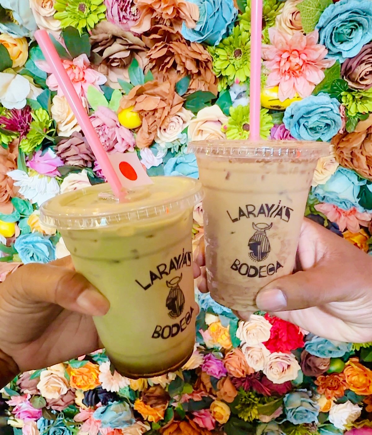 Two hands hold plastic cups of iced drinks with pink straws labeled “Larayia’s Bodega” in front of a colorful wall covered in vibrant artificial flowers.