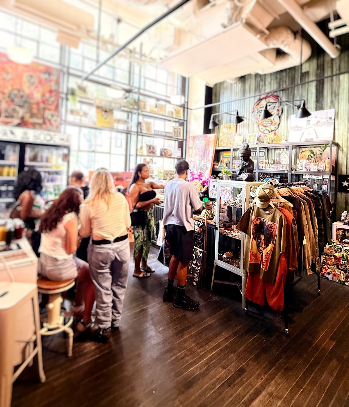 People browse clothes, art, and gifts inside LaRayia’s Bodega with large windows, shelves, and wall art. Some stand at a counter while others look at items on racks and displays.
