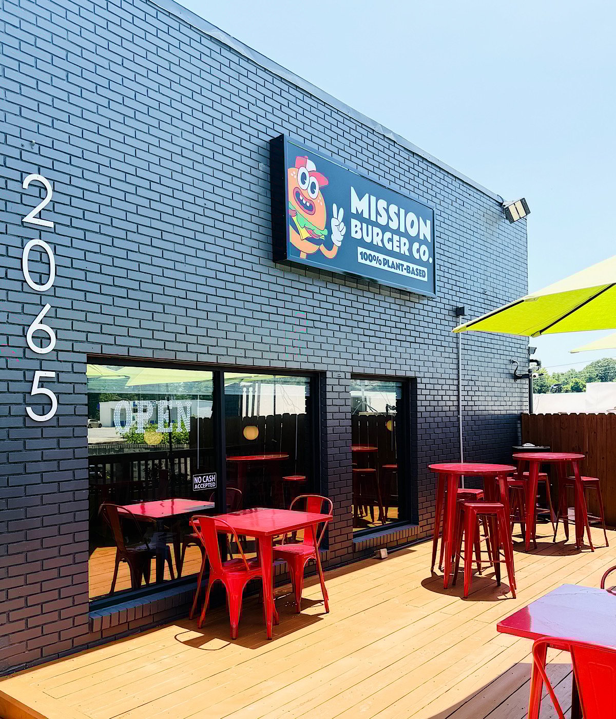 Outdoor patio of Mission Burger Co. restaurant with bright red tables and chairs, a large OPEN sign, and a colorful burger logo on the gray brick wall. A lime green umbrella provides shade on a sunny day.