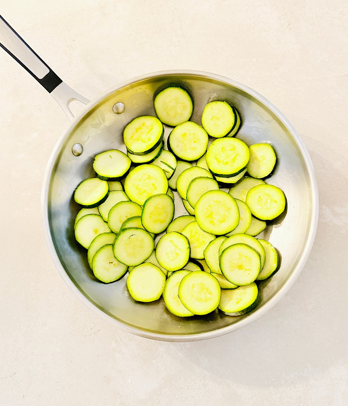 A stainless steel pan filled with sliced zucchini for Vegan Zucchini Quiche sits on a light-colored surface. The zucchini slices are arranged in a single layer inside the pan.