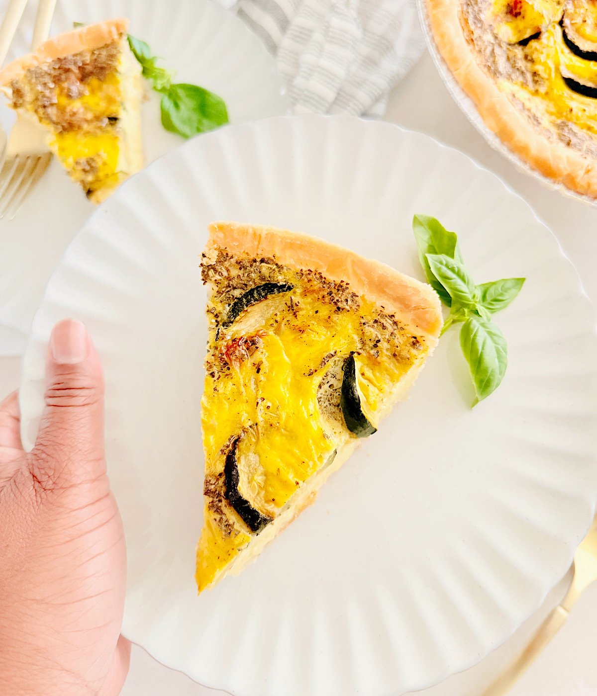 A hand holds a white plate with a slice of Vegan Zucchini Quiche garnished with fresh basil; another plate and the remaining quiche are visible in the background.
