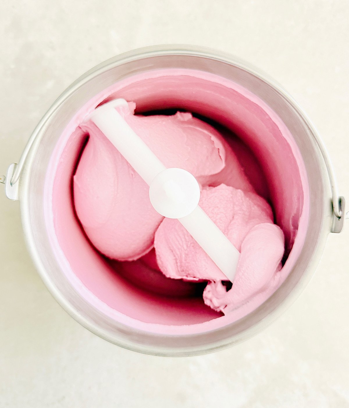 A metal ice cream maker bowl filled with smooth, pink ice cream, shown from above with the churning paddle in the center.