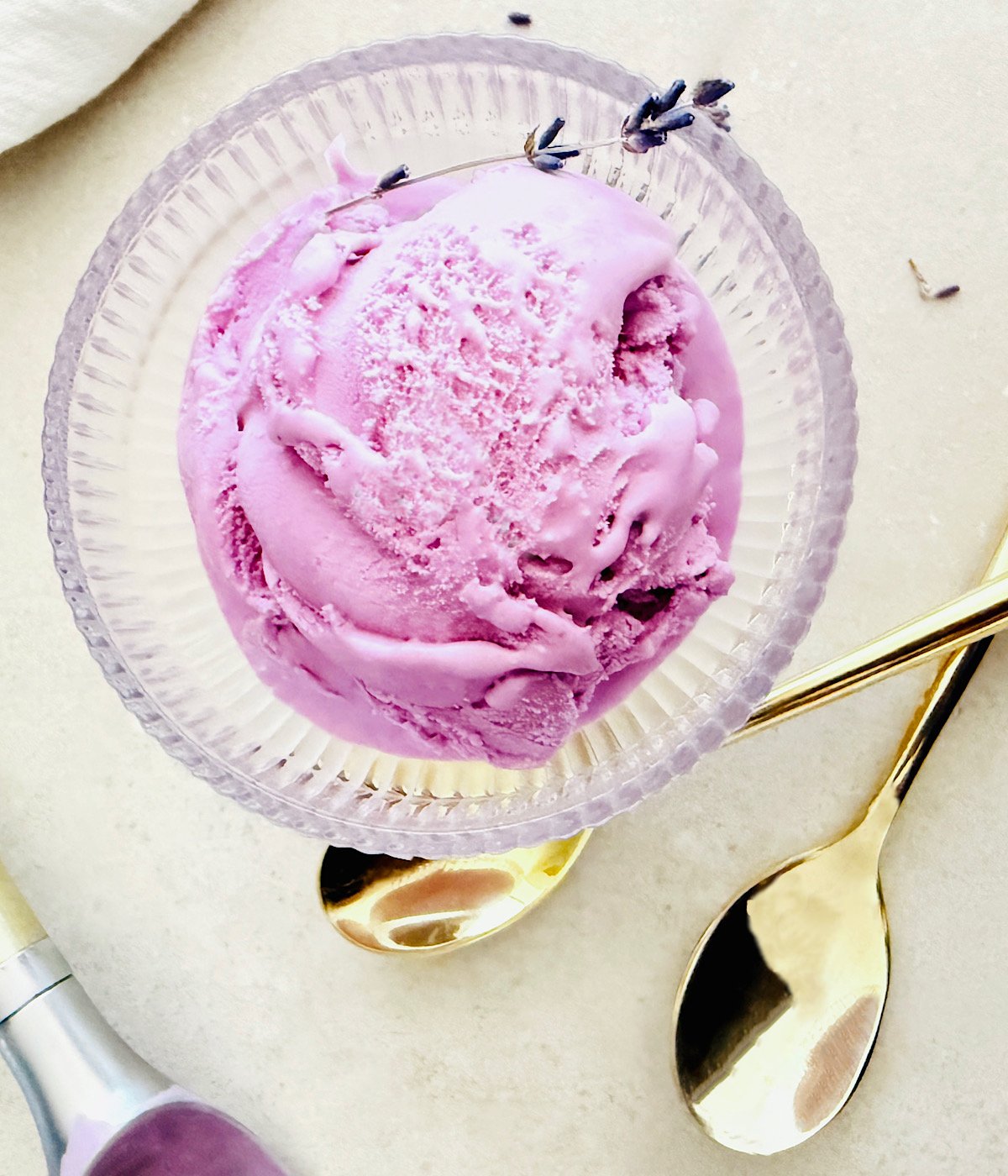 A glass bowl holds a scoop of bright purple lavender ice cream, garnished with sprigs of lavender. Two shiny gold spoons and an ice cream scoop are nearby on a light surface.
