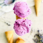 A close-up of a double scoop of bright purple ice cream in a waffle cone, with more cones and scattered lavender sprigs blurred in the background.