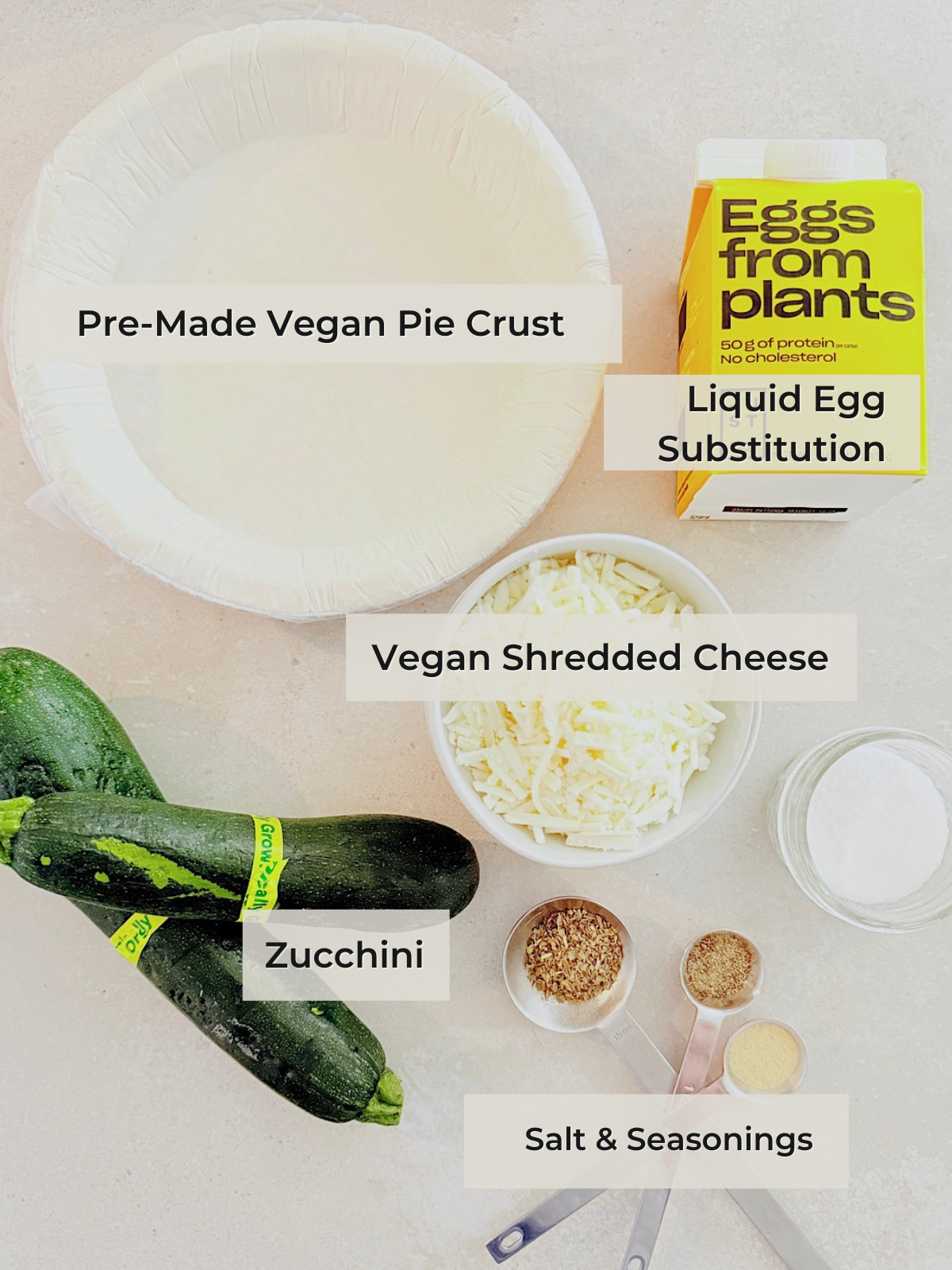Overhead view of ingredients for a vegan quiche: premade vegan pie crust, liquid egg substitute, vegan shredded cheese, two zucchinis, salt, and seasonings, all labeled on a light countertop.