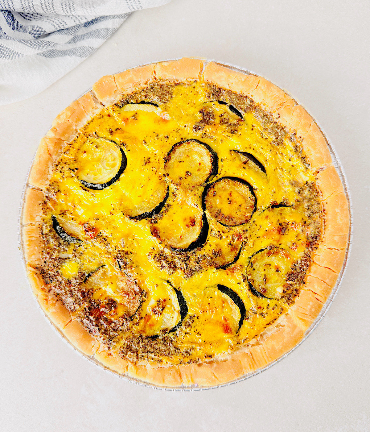 A Vegan Zucchini Quiche in a pie crust, filled with visible slices of zucchini and a yellow egg mixture, sits on a light surface with a striped kitchen towel partially visible in the corner.