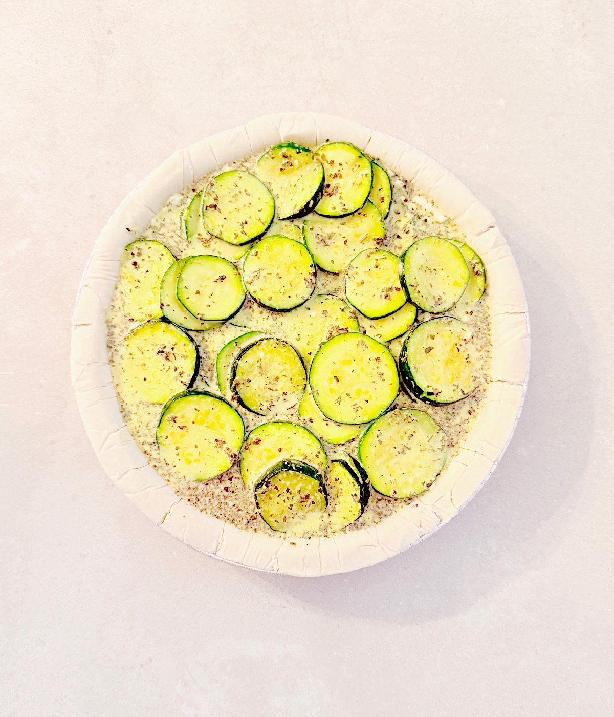A round, unbaked pie crust filled with overlapping slices of zucchini, sprinkled with herbs and seasoning, is ready to become a delicious Vegan Zucchini Quiche on a light-colored surface.