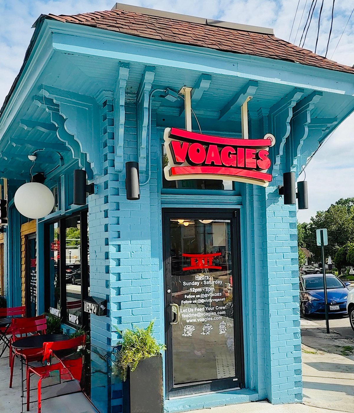 A blue brick restaurant with a red and white VOAGIES sign above the glass door. Red chairs and tables sit outside, and the door displays the restaurants hours and website. A large round lamp hangs near the entrance.