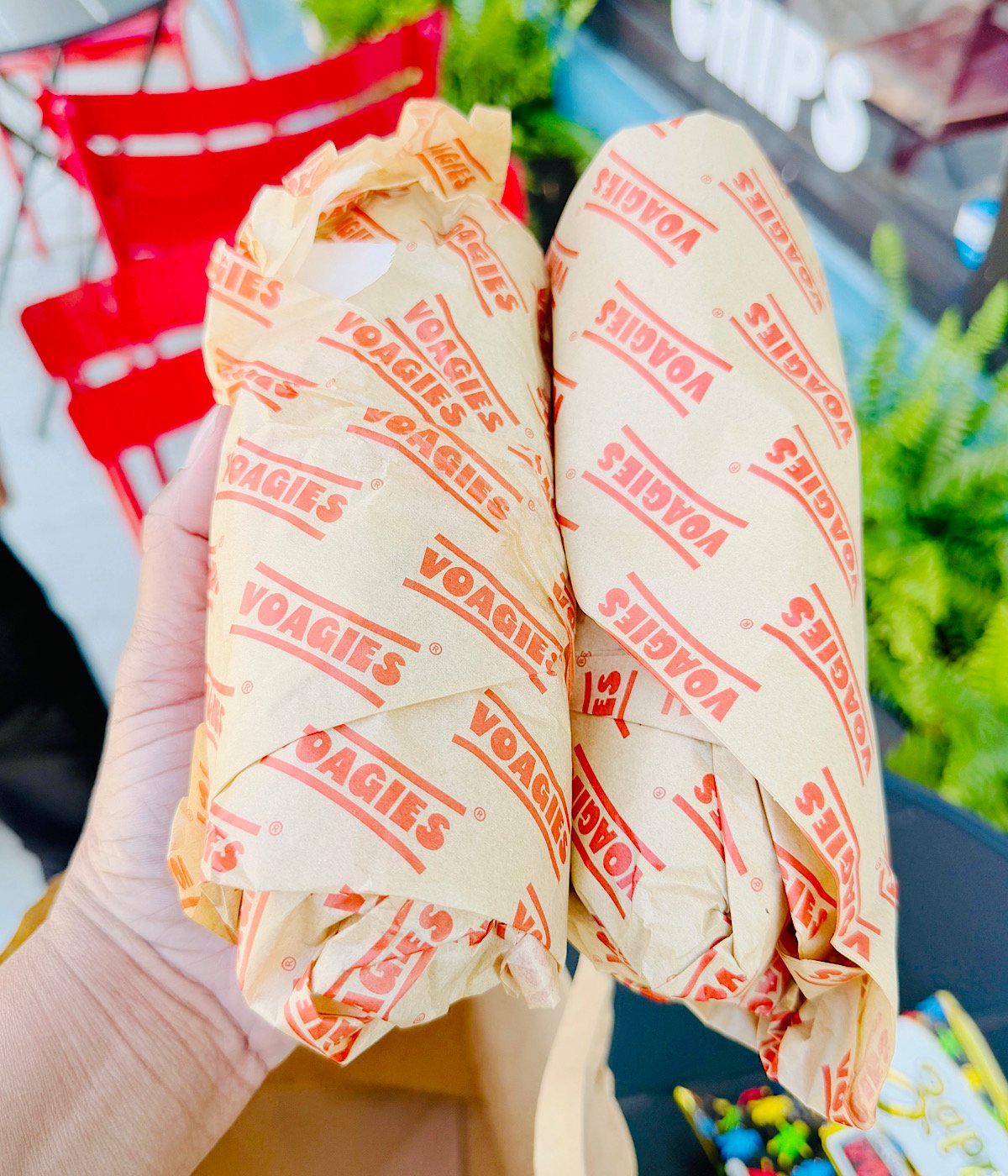 A hand holds two wrapped hoagie sandwiches in yellow paper labeled VOAGIES. Red chairs and green plants are visible in the background.