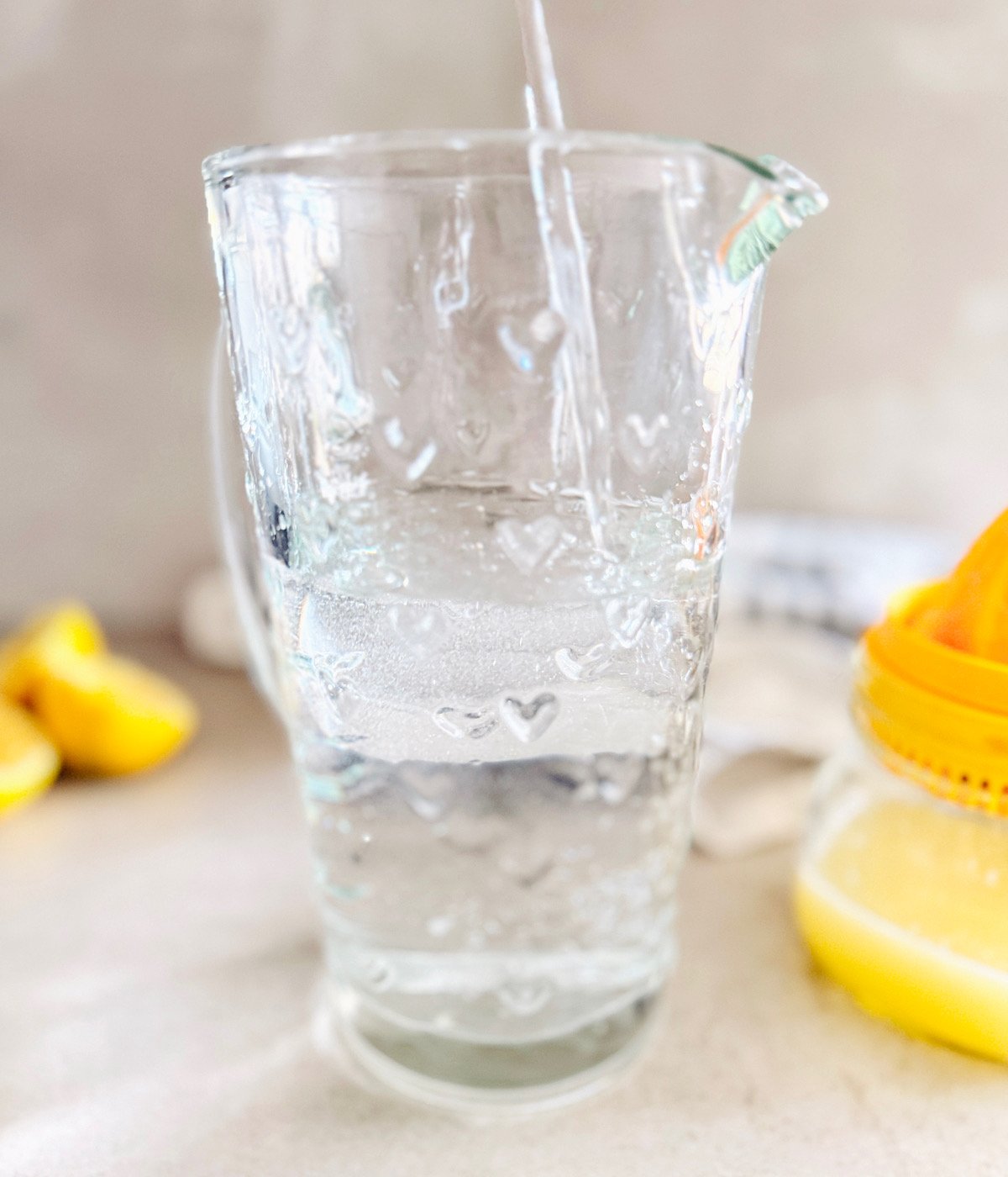 A clear glass pitcher being filled with water. In the background, there are lemon halves and a yellow citrus juicer. The scene is brightly lit with a clean, soft focus.