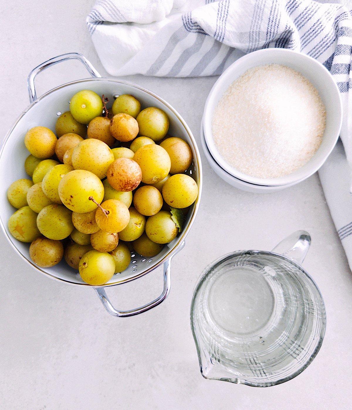 A colander filled with yellow-green muscadine, a bowl of granulated sugar, and a glass pitcher of water sit on a light surface next to a white and blue striped kitchen towel.