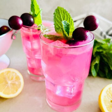Two glasses of bright pink Muscadine Lemonade with ice, garnished with mint leaves and muscadine on skewers. A lemon half and fresh mint are nearby on the countertop.