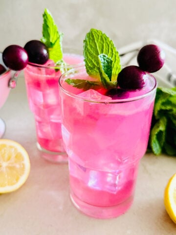 Two glasses of bright pink Muscadine Lemonade with ice, garnished with mint leaves and muscadine on skewers. A lemon half and fresh mint are nearby on the countertop.