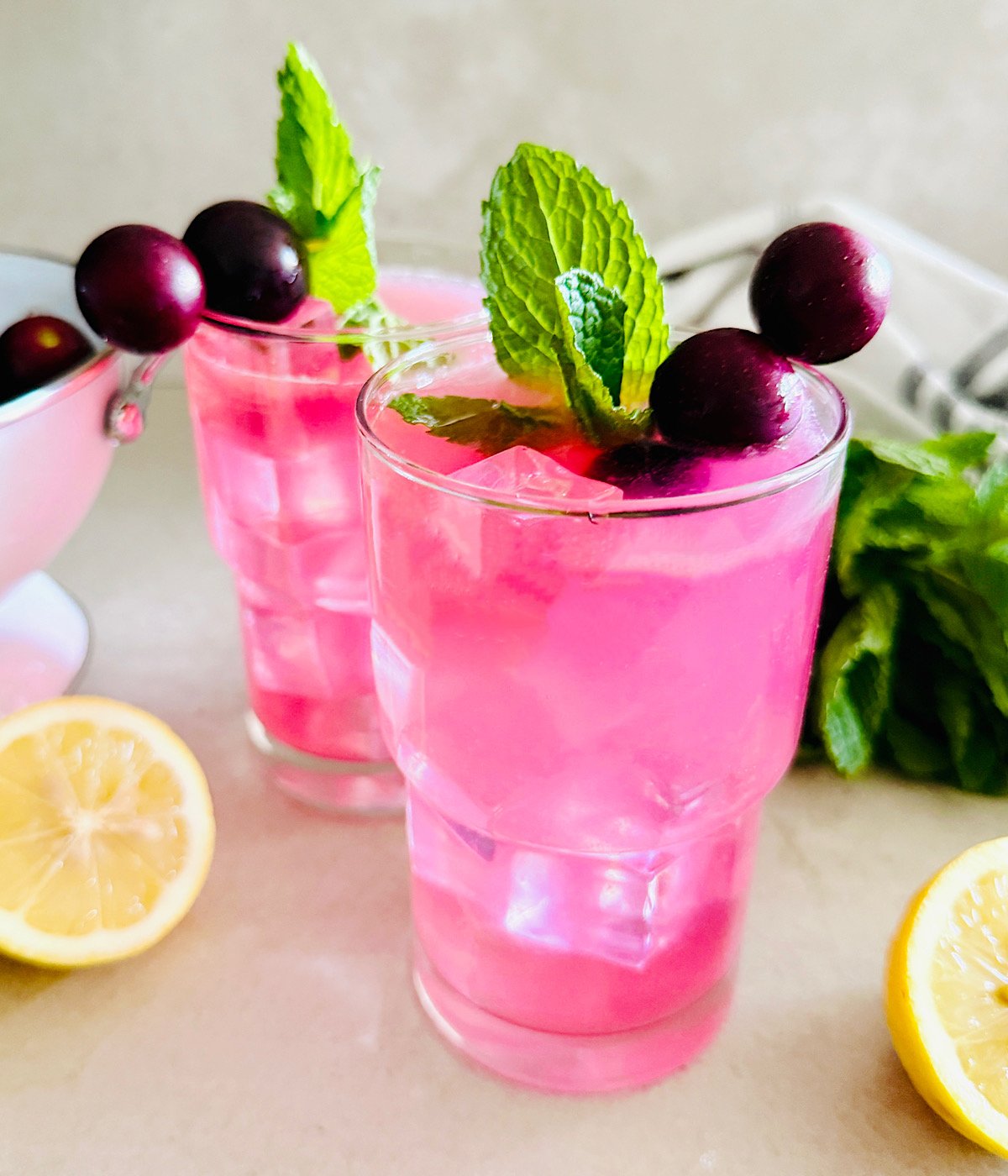 Two glasses of bright pink Muscadine Lemonade with ice, garnished with mint leaves and muscadine on skewers. A lemon half and fresh mint are nearby on the countertop.
