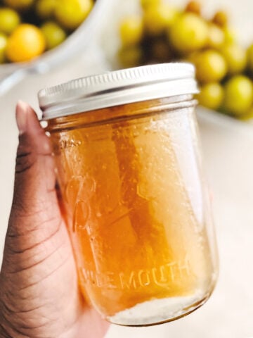 A hand holds a clear glass jar filled with muscadine simple syrup; blurred green and yellow muscadine are visible in bowls in the background.
