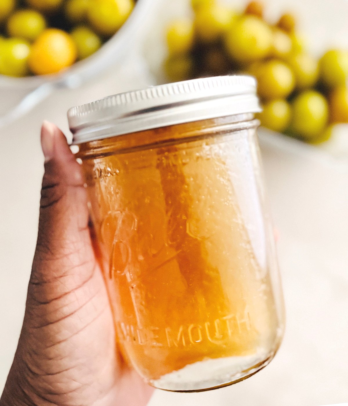 A hand holds a clear glass jar filled with muscadine simple syrup; blurred green and yellow muscadine are visible in bowls in the background.