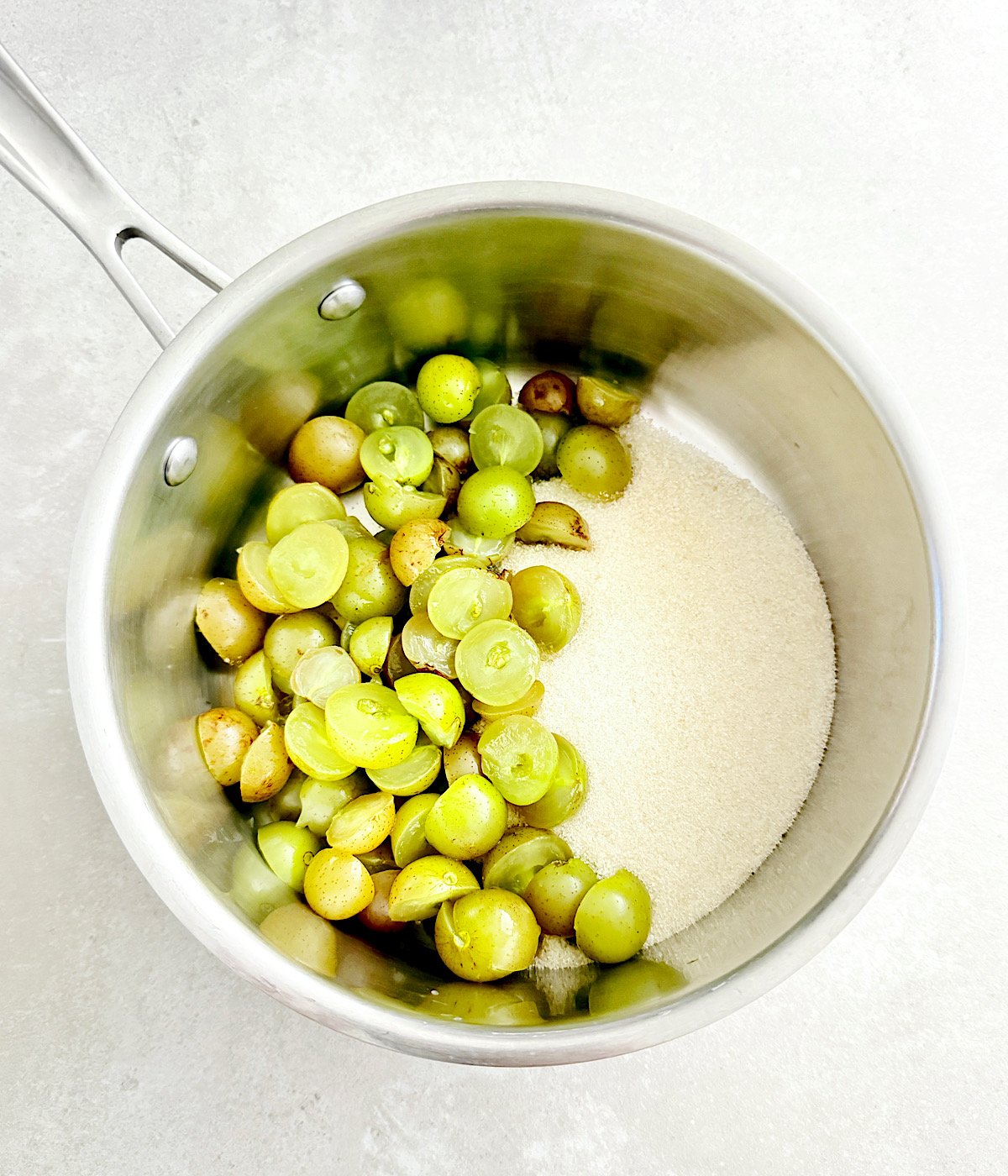 A stainless steel saucepan containing halved green muscadine and granulated sugar on a light countertop.