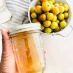 A hand holds a mason jar filled with light golden simple syrup, while a white colander with round yellow and green fruit sits in the background on a light surface.