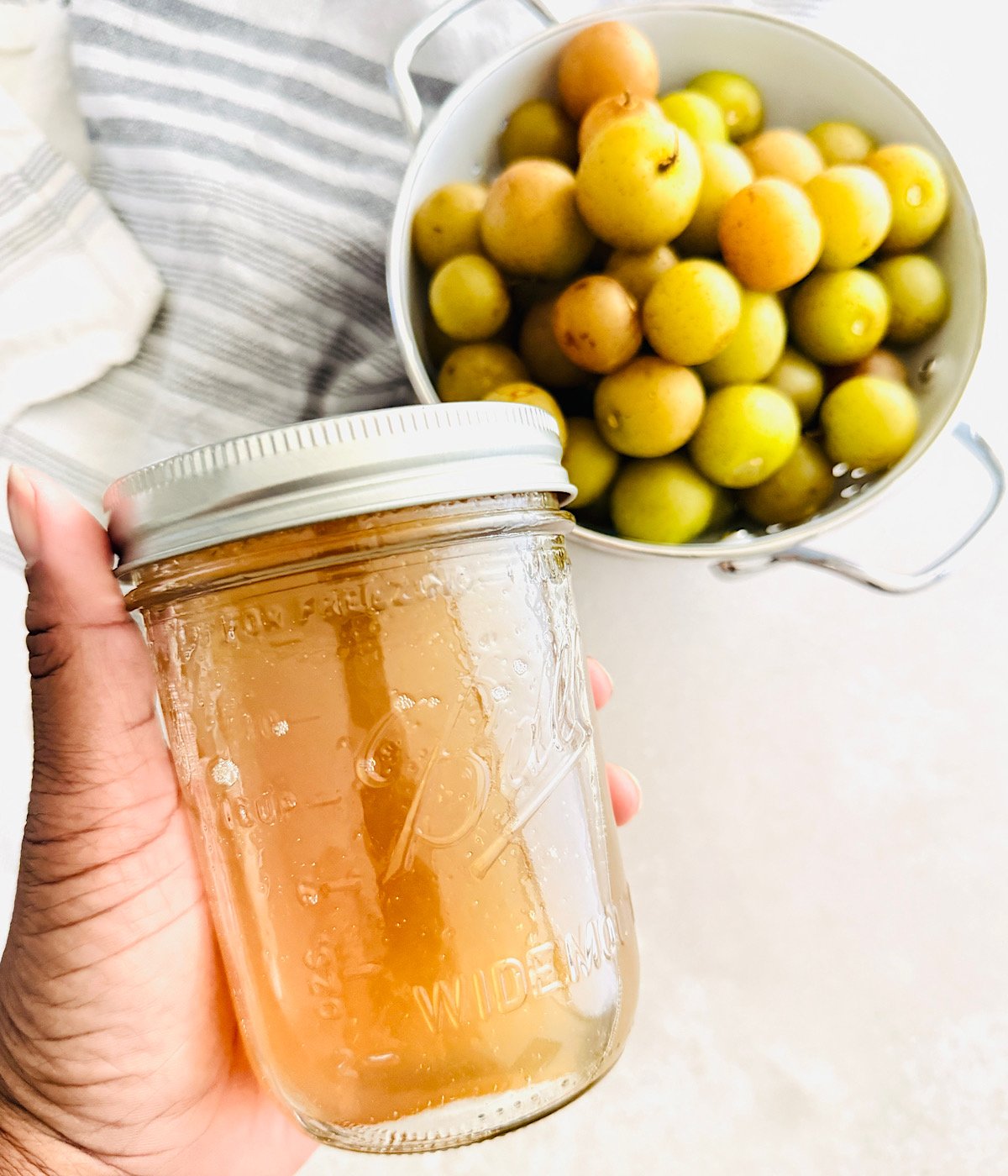 A hand holds a mason jar filled with light golden simple syrup, while a white colander with round yellow and green fruit sits in the background on a light surface.