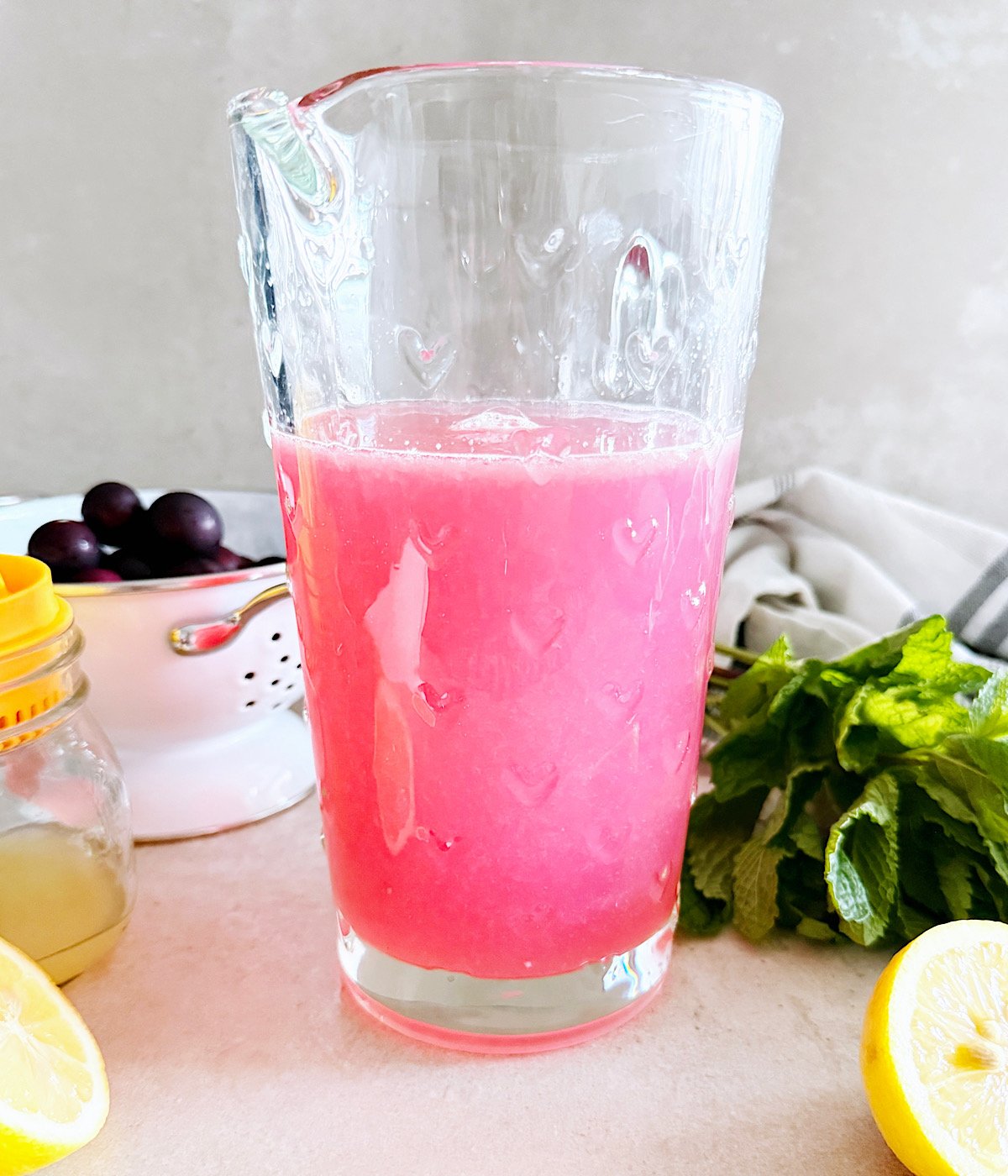A tall glass pitcher filled with bright pink lemonade sits on a counter, surrounded by fresh mint leaves, a sliced lemon, a jar of lemon juice and a bowl of dark muscadine in the background.