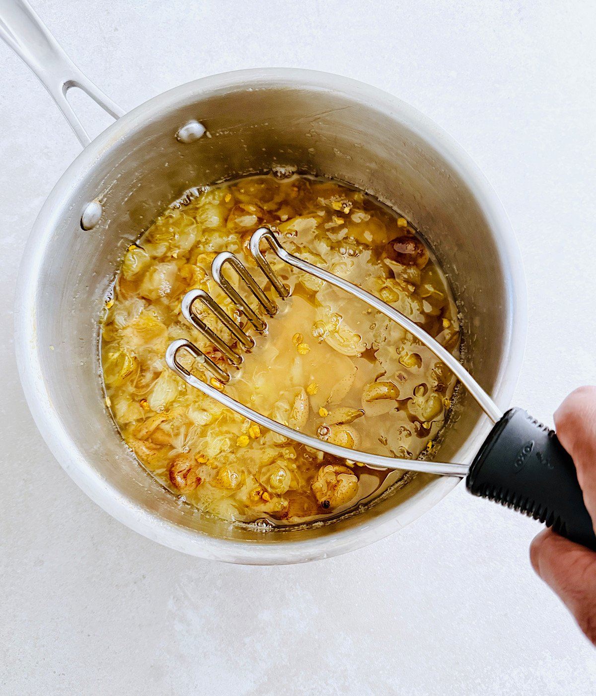 A hand holds a potato masher over a saucepan filled with cooked, diced muscadine, preparing to mash the mixture.