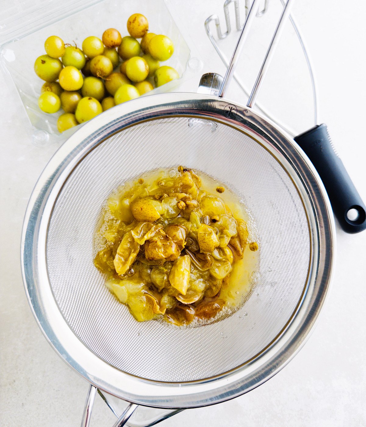 A metal strainer holds mashed yellow-green muscadines and juice above a bowl. A potato masher rests on the strainer. A plastic container of whole, round yellow-green fruits sits nearby on a white surface.
