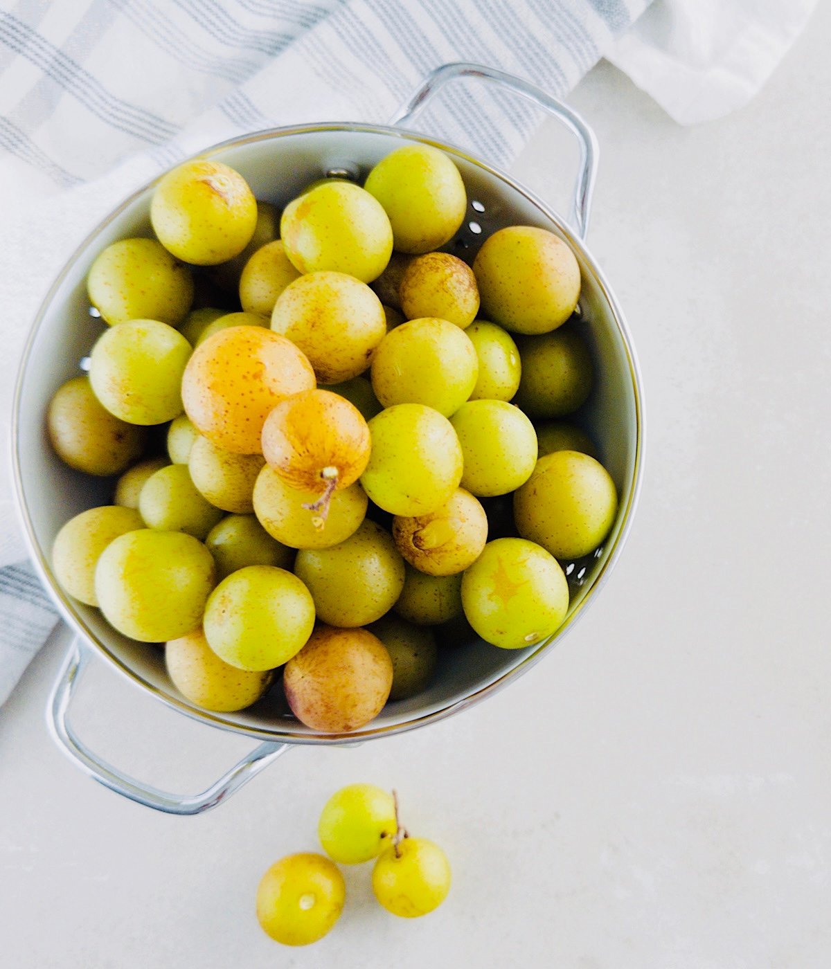A metal colander filled with fresh yellow-green muscadine grapes sits on a white surface, with a few grapes lying beside it and a striped kitchen towel in the background.