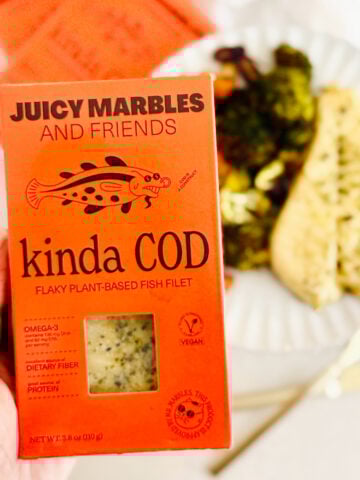A hand holds a box of Vegan Juicy Marbles “Kinda Cod” flaky plant-based fish filet in front of a plate with vegan fish and vegetables. The bright orange packaging highlights its vegan and protein content.