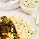 A plate with baked vegan fish covered in a creamy vegan fish dill sauce, served with roasted broccoli and carrots. In the background, a glass sauce boat with more of the vegan fish dill sauce sits next to a fork.
