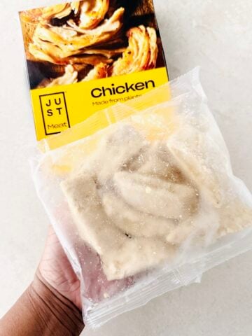 A hand holds a package of plant-based Just Meat Chicken, showing the frozen contents in a clear plastic bag in front of the products yellow and black box.