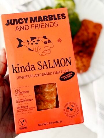 A hand holds a box of Juicy Marbles Kinda Salmon, a plant-based fish fillet. The orange packaging highlights protein, fiber, omega-3, and vegan labels, with food blurred on a plate in the background.