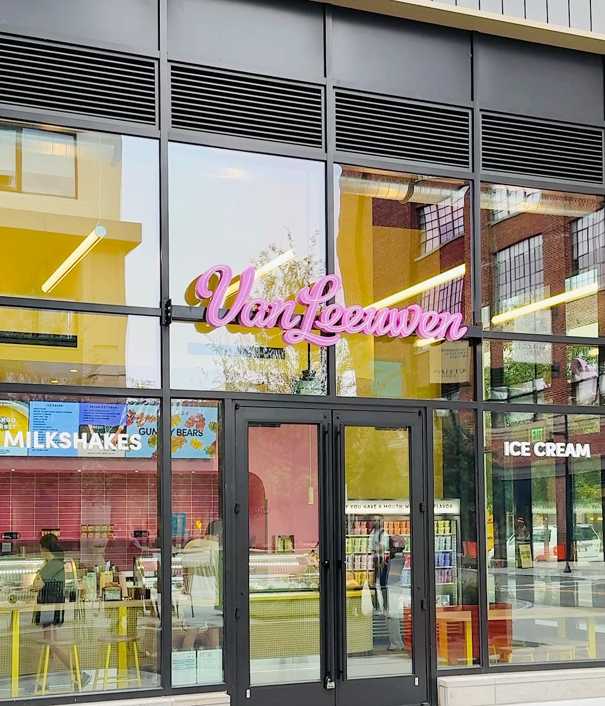The storefront of Van Leeuwen, featuring large windows, a pink neon sign, and visible interior displays for milkshakes and ice cream. The reflection of nearby buildings appears on the glass.