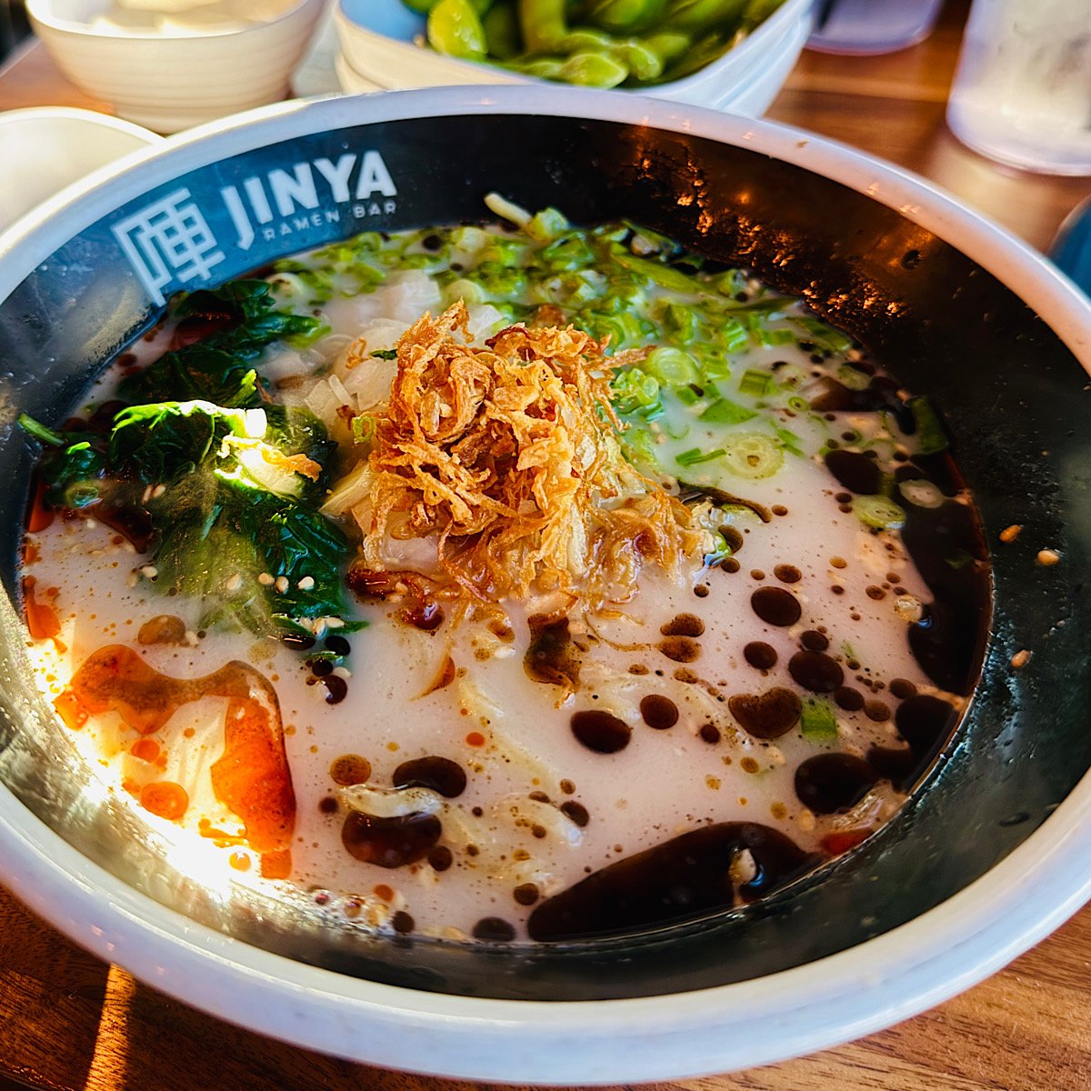 A bowl of ramen from Jinya Ramen Bar, topped with sliced green onions, spinach, crispy fried onions, and black garlic oil in a creamy broth, with small dishes in the background.